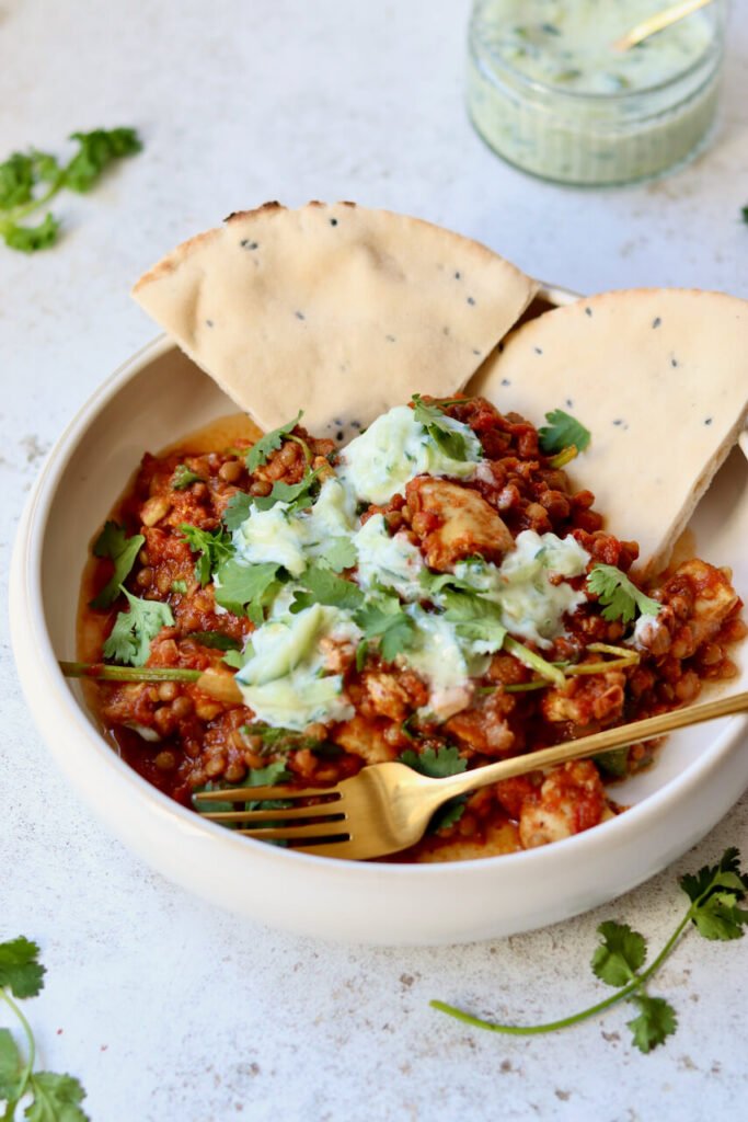 Healthy one-pot chicken dhansak served in a white bowl with lentils, fresh coriander, cucumber raita and seeded flatbread — a high-protein, lighter take on the fakeaway curry classic.