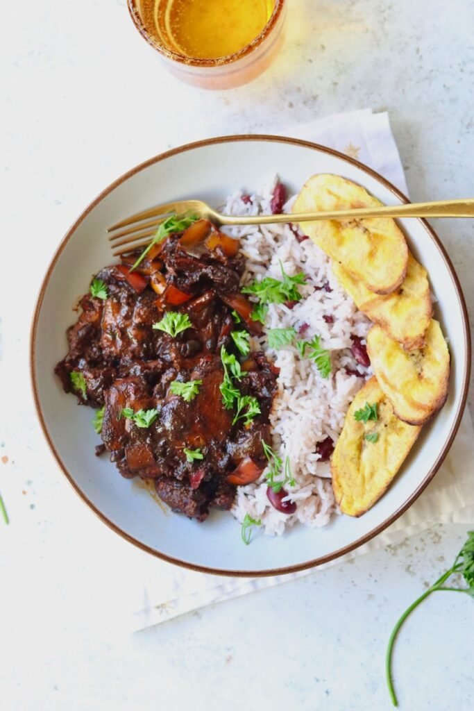 Brown stew chicken plated with rice and peas, plantain and a gold fork