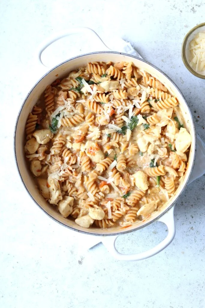 One-pot high-protein Tuscan chicken pasta in a white casserole pot, with chickpea rotini, wilted spinach and parmesan shavings