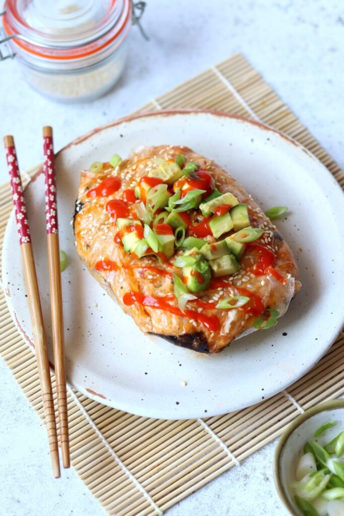 Rice paper sushi bake topped with diced avocado, spring onion and sriracha on a speckled plate with chopsticks
