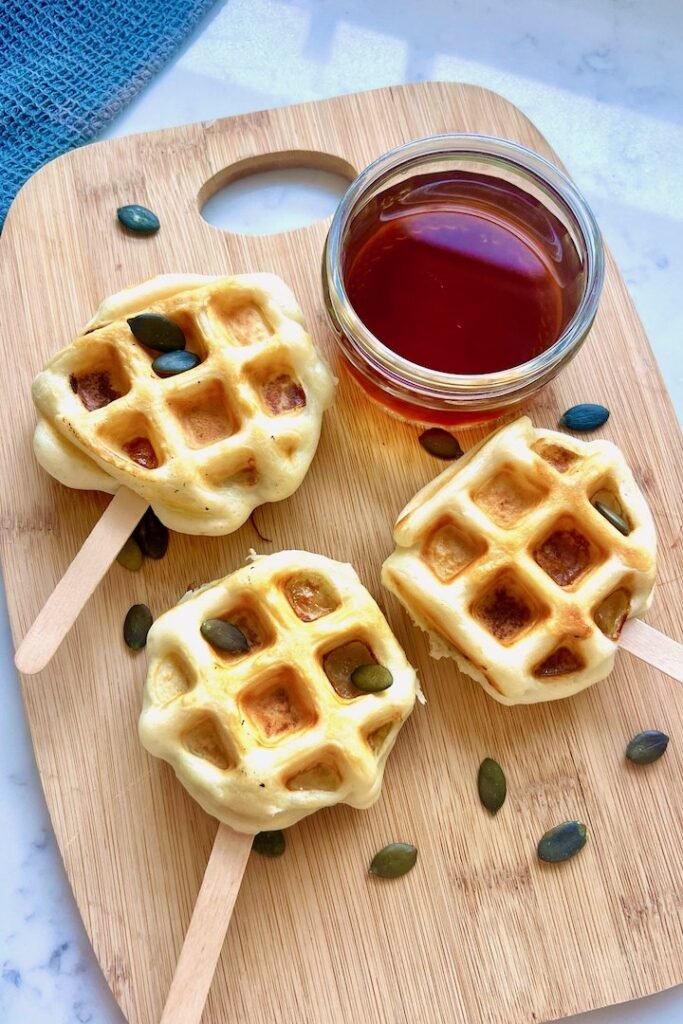 Three apple waffle pops on a wooden board with a jar of maple syrup