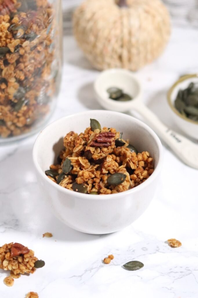 Bowl of pumpkin pie protein granola with a gold spoon, small pumpkins and pumpkin seeds beside it