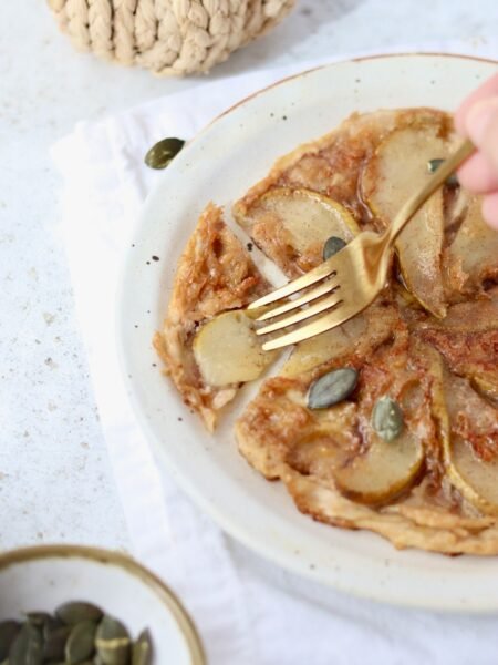 Rice paper tarte tatin on a plate with pear slices fanned out and a fork lifting a bite, pumpkin seeds scattered alongside