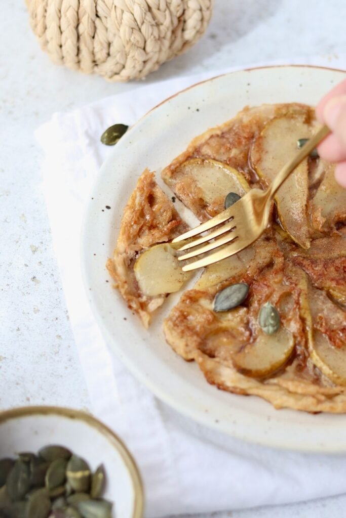 Rice paper tarte tatin on a plate with pear slices fanned out and a fork lifting a bite, pumpkin seeds scattered alongside
