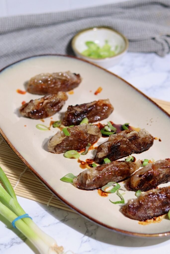 A platter of pan fried duck dumplings wrapped in rice paper, topped with sliced spring onion